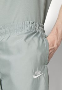 Ljust grå idrottsshorts tillverkade av ett glatt tyg med resårmidja, sidofickor och en vit broderad Nike-logga.