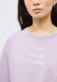 Νεαρή γυναίκα με σκούρα μαλλιά που φοράει μωβ φούτερ με λευκό κείμενο που λέει «ΤΟ ΜΕΛΛΟΝ ΕΙΝΑΙ ΔΙΚΟ ΜΑΣ» και μικρά σκουλαρίκια κρίκους.