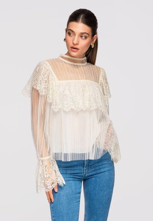 Kvinde iført gennemsigtig hvid bluse i blonder med flæser og høj krave, kombineret med blå jeans, der kigger mod venstre mod en lys baggrund.