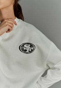 Valkoinen huppari, jossa on pyöreä graafinen logo, jossa lukee "SF" mustalla. Pehmeä kangas, rento istuvuus ja ribbireuna.
