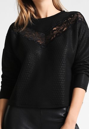 Kvinde iført en sort, langærmet tekstureret top med en V-formet sort blonde detalje ved udskæringen, kombineret med sorte bukser.