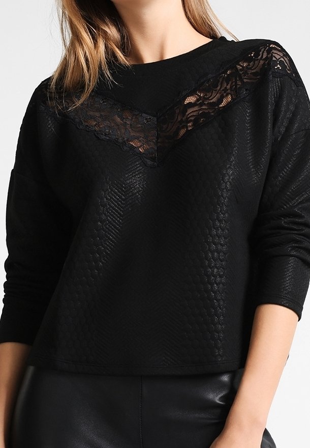 Femme portant un haut texturé noir à manches longues avec un détail en dentelle noire en forme de V au niveau de l'encolure, associé à un pantalon noir.