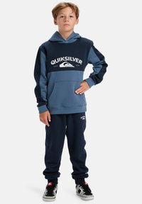 Navy and blue color-blocked hoodie with front pocket, featuring white "Quiksilver" logo. Matched with dark jogger pants and black sneakers.