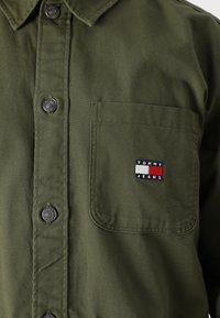 Chemise olive boutonnière avec une poche poitrine, dotée de boutons noirs et d'une patch logo multicolore de Tommy Jeans sur la poche.