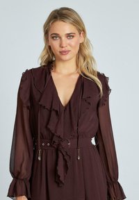 Bordeauxklänning med volangdetaljer, långa genomskinliga ärmar, djup V-ringning och ett bälte i midjan med dekorativa hårdvarudetaljer.