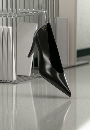 Svart høyhælte sko med spiss tå og gyllent logo, plassert på en reflekterende overflate nær en kromtråd-filholder med dokumenter.