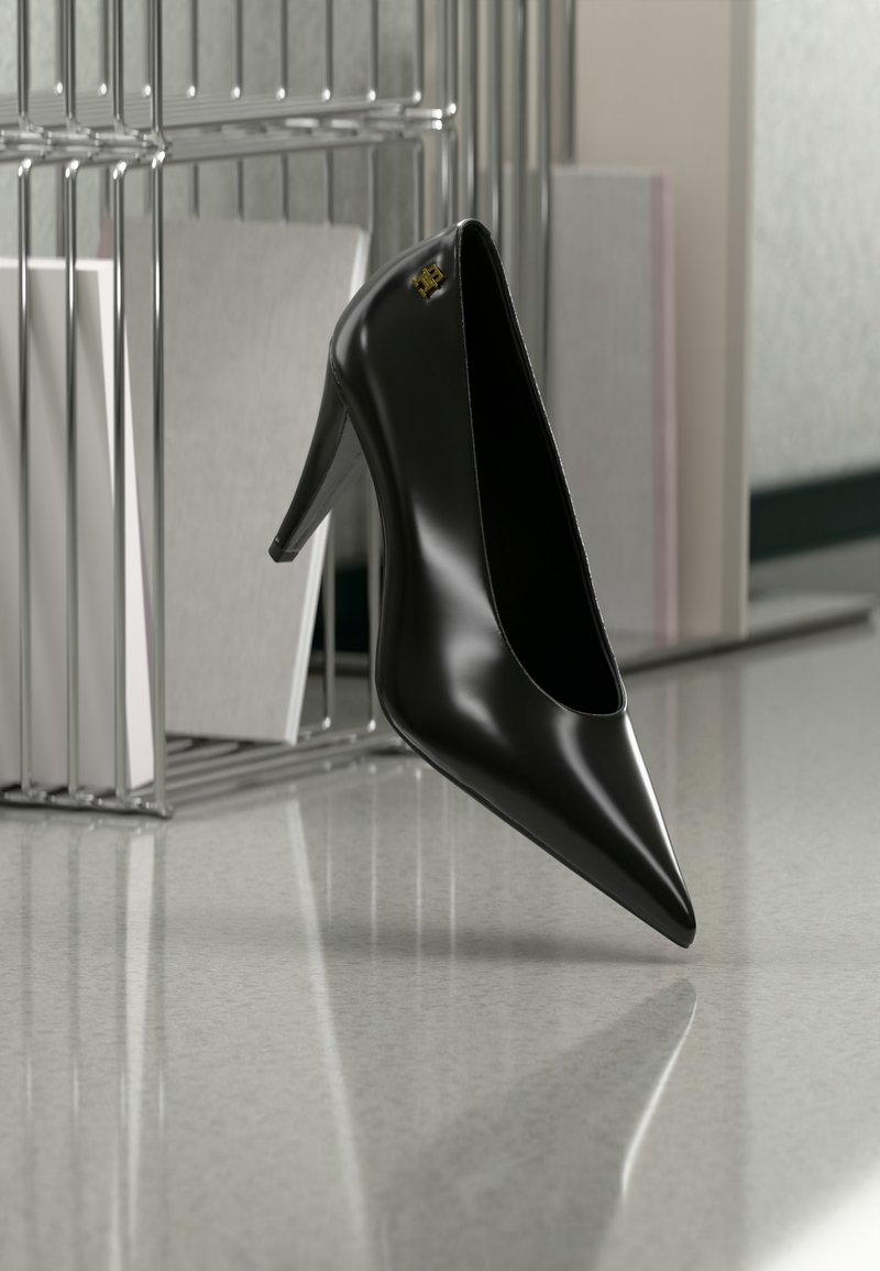 Μαύρο γυναικείο παπούτσι με ψηλό τακούνι και μυτερή μύτη με χρυσό λογότυπο, τοποθετημένο σε μια ανακλαστική επιφάνεια κοντά σε χρωμιωμένο μεταλλικό οργανωτή αρχείων με έγγραφα.