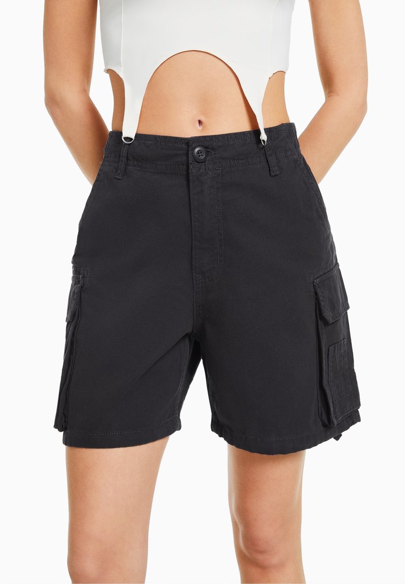 Top court blanc avec détails découpés porté avec un short cargo noir mi-cuisse avec poches à rabat et fermeture par boutons.
