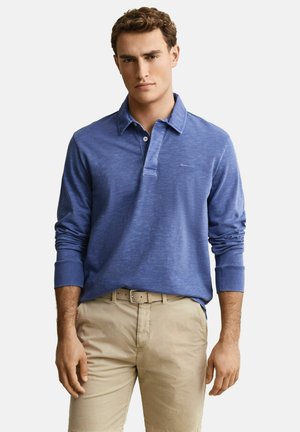 Jeune homme portant un polo bleu à manches longues, un pantalon beige et une ceinture, debout devant un fond clair uni.