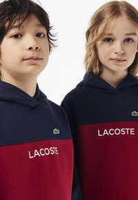 Due bambini che indossano felpe con cappuccio Lacoste. Le felpe presentano blocchi di colore blu navy e rosso, un logo del coccodrillo verde e testo bianco con la scritta "LACOSTE".