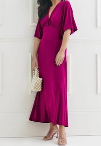 Robe maxi en jersey fuchsia avec un décolleté en V profond et des manches chauve-souris, associée à un sac à main en perles brodées et des sandales à talons avec lanières.