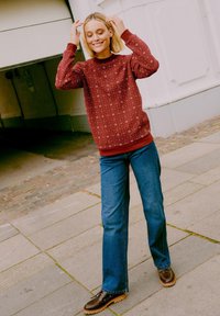 Sudadera roja con estampado, con un cuello redondo, combinada con jeans rectos azules y zapatos marrones de cordones sobre una superficie pavimentada.