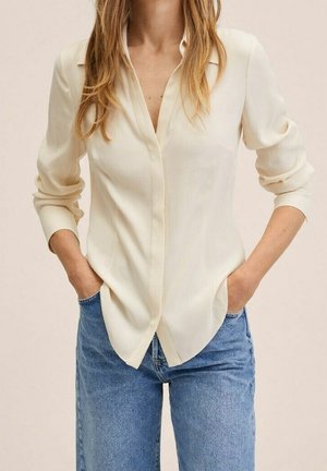 Vrouw draagt een crèmekleurige blouse met lange mouwen die zijn opgerold en een blauwe spijkerbroek, handen in de voorzakken.