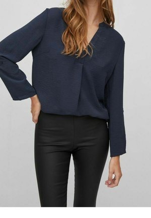 Marineblauwe blouse van lichtgewicht textuurstof met een V-hals, lange mouwen en een zak. Gecombineerd met aansluitende zwarte leren-achtige pants.