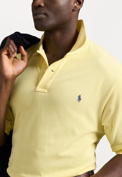 Polo amarelo feito de tecido texturizado, com colarinho clássico, pala de três botões e um pequeno logótipo azul no lado esquerdo do peito.