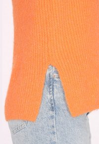Oranger gestrickter Pullover mit einem gerippten Muster, der einen seitlichen Schlitz aufweist. Kombiniert mit hellblauen Jeans.
