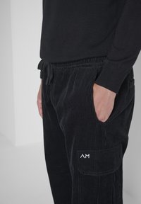 Schwarze Cordhosen mit einem Kordelzugbund, seitlichen Taschen und einer aufgesetzten Tasche mit einem silbernen Logo am linken Bein.