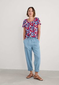 Top de manga corta multicolor con patrones abstractos en rojo, azul y blanco. Pantalones de tiro medio en azul claro con bolsillos y ajuste casual. Sandalias marrones.