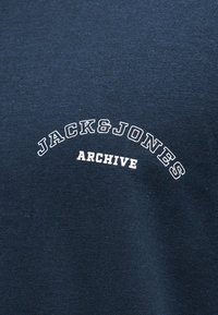 Ύφασμα σε ναυτικό μπλε χρώμα με λευκό κείμενο "JACK & JONES" καμπυλωμένο πάνω από τη λέξη "ARCHIVE" σε κεφαλαία γράμματα στο κέντρο.