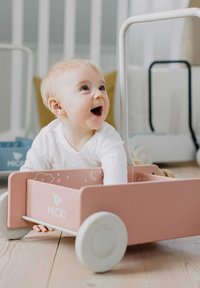 Trælegetøjsvogn i pink med hvide hjul. Har legende sky- og måne designs indeni. Barnet leger med vognen og smiler.