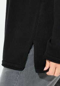 Maglione nero lavorato a maglia con una texture a costine, fessure laterali e polsini arrotolati, abbinato a jeans grigi. Inquadratura ravvicinata di una mano che tiene l'orlo.