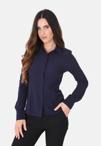 Camicia a maniche lunghe blu navy con bottoni, dal design aderente, texture liscia e colletto a punta, indossata con pantaloni neri.