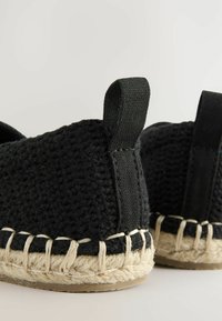 Schwarze gewobene Espadrilles mit einer strukturierten Oberseite, ausgestattet mit zwei Zuglaschen und einer Juteschnur-Sohle, verziert mit beigefarbenen Nähten.