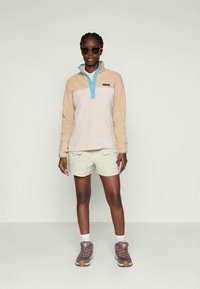 Lichtbeige fleece pullover met een turquoise kraag, gecombineerd met lichtgroene shorts en bruine wandelschoenen, voorzien van een logo patch.