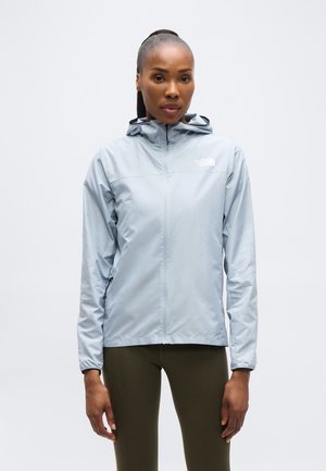 Kvinde iført en lyseblå hættetrøjejakke fra North Face og olivengrønne leggings, stående mod en ensfarvet hvid baggrund.