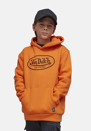 Garçon portant une casquette noire et un sweat à capuche orange Von Dutch, les mains dans la poche avant, debout devant un fond clair uni.