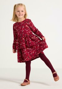 Czerwona kwiecista sukienka z długimi rękawami i warstwową spódnicą, zestawiona z bordowymi rajstopami prążkowanymi oraz jasnoróżowymi butami z falbaniastymi krawędziami.