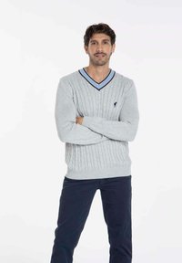 Pull en maille torsadée gris clair avec un col en V et des accents bleu foncé. Porté sur un pantalon sombre, présentant un logo sur le côté gauche.