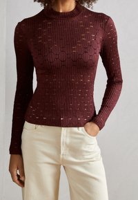 Burgundy ribbad långärmad topp med texturmönster, rund hals, figurnära silhuett, bärs med krämfärgade byxor, hand i fickan.