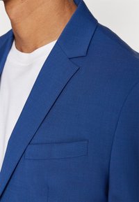 Blå blazer med strukturerad tyg, spetsiga slag och en enda bröstficka, kombinerad med en vit t-shirt under.