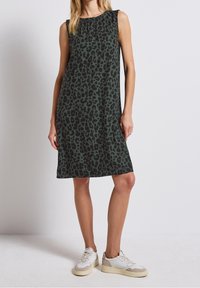 Žena v bezrukávových tmavě zelených šatech ke kolenům s černým leopardím vzorem a bílými teniskami stojí proti jednoduchému pozadí.