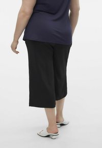 Czarne spodnie culotte z miękkiego materiału, o luźnym kroju, opadające poniżej kolana, w połączeniu z białymi sandałami na obcasie z odkrytymi palcami.