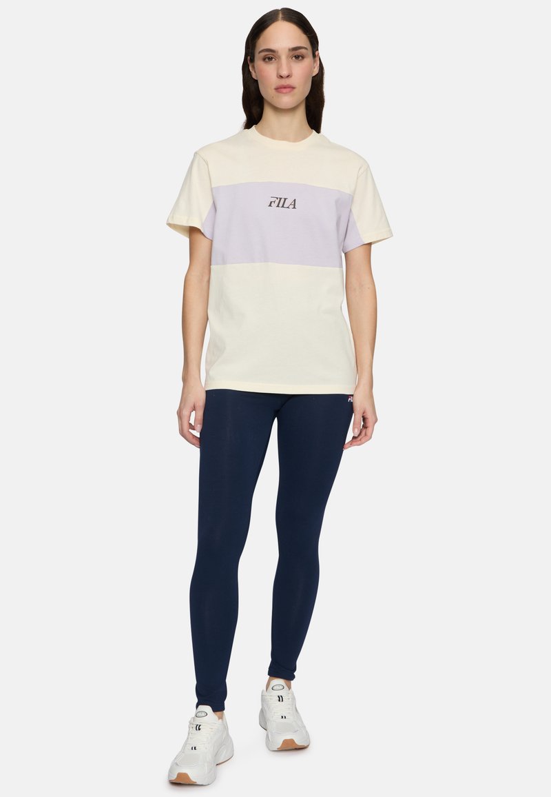 T-shirt en coton de couleur jaune pâle et blocs de lavande avec le logo "FILA", associé à des leggings navy et des sneakers blanches avec des semelles marron.