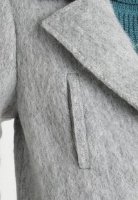 Cappotto di lana grigio con una finitura morbida e testurizzata. Presenta un colletto a risvolto e una tasca verticale sul lato anteriore.