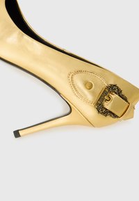 Gold leather high-heeled pump featuring a pointed toe, sleek silhouette, and an ornate metal buckle accent on the heel.