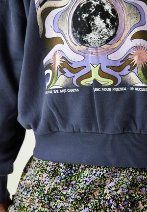Sweatshirt navy avec un design graphique coloré comprenant des motifs célestes et du texte. Poignets et ourlet côtelés, associé à une jupe à motifs floraux.