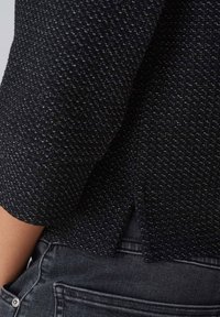 Schwarzer strukturierter Pullover mit silbernen Flecken, mit einem geteilten Saum, kombiniert mit dunkelgrauen Jeans. Der Stoff hat ein leicht erhabenes Muster.