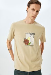 Beige bomulds t-shirt med et trykt design af et granatæble, en pære og en kiwi med tekst. Korte ærmer, rund hals, afslappet pasform.