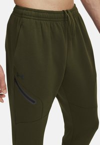 Olive green joggers with an elastic waistband, zippered left pocket, and subtle black logo detail on the thigh. Smooth, athletic fabric.