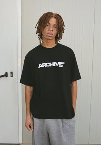 Černé bavlněné tričko s potiskem "ARCHIVE INC" v tučných bílých písmenech, spárované se šedými šortkami. Uvolněný střih s krátkými rukávy.
