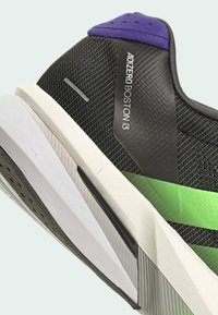 Baskets de sport noires avec une tige texturée, ornées d'une bande d'accent verte et d'un talon violet. La semelle est blanche avec un motif unique.