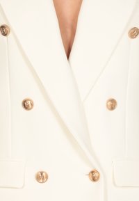 Blazer doppiopetto bianco con bottoni dorati, caratterizzato da un profondo scollo a V e spalle strutturate, realizzato in tessuto liscio.