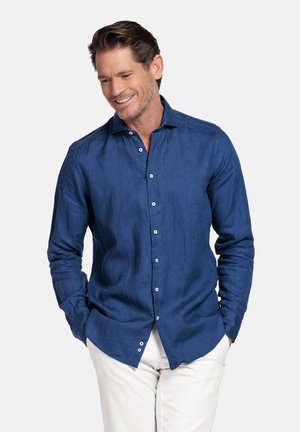 Man die lacht, gekleed in een blauw overhemd met lange mouwen en witte broek, met handen in de zakken, tegen een effen lichte achtergrond.