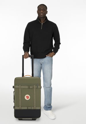 Olive green wheeled suitcase with black accent, zipper closure, and decorative straps. Sturdy handle and logo patch visible.