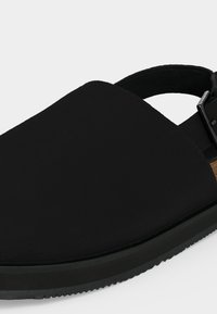 Μαύρο παπούτσι χωρίς κορδόνια με κλειστή μύτη, ρυθμιζόμενο λουράκι στη φτέρνα με αγκράφα και υφή στη σόλα για καλύτερη πρόσφυση.