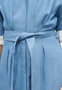 Φόρεμα γαλάζιο με ζώνη από ύφασμα δεμένη στη μέση, με πιέτες και μανίκια γυρισμένα για να φαίνεται ένα λευκό εσωτερικό στρώμα.